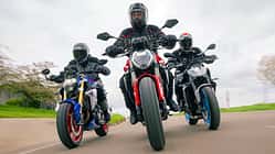 Das Foto zeigt drei Naked Bikes in enger Kurvenfahrt: Ducatti Monster, BMW F900 R und Yamaha MT-09. Die Fahrer in vollständiger Schutzkleidung bewegen die Motorräder mit deutlicher Schräglage auf einer kurvigen Landstraße, Front- und Fahrwerksdetails sind klar erkennbar. Die Szene wirkt wie eine Gruppen- bzw. Vergleichsfahrt unter trockenen Bedingungen.