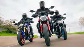 Das Foto zeigt drei Naked Bikes in enger Kurvenfahrt: Ducatti Monster, BMW F900 R und Yamaha MT-09. Die Fahrer in vollständiger Schutzkleidung bewegen die Motorräder mit deutlicher Schräglage auf einer kurvigen Landstraße, Front- und Fahrwerksdetails sind klar erkennbar. Die Szene wirkt wie eine Gruppen- bzw. Vergleichsfahrt unter trockenen Bedingungen.