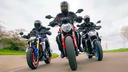 Ducatti Monster, BMW F900 R, Yamaha MT-09, Ducati Monster, Naked Bike, Motorradvergleich, Kurvenfahrt, Landstraße, Gruppenfahrt, Motorradtest Das Foto zeigt drei Naked Bikes in enger Kurvenfahrt: Ducatti Monster, BMW F900 R und Yamaha MT-09. Die Fahrer in vollständiger Schutzkleidung bewegen die Motorräder mit deutlicher Schräglage auf einer kurvigen Landstraße, Front- und Fahrwerksdetails sind klar erkennbar. Die Szene wirkt wie eine Gruppen- bzw. Vergleichsfahrt unter trockenen Bedingungen.