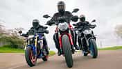 Das Foto zeigt drei Naked Bikes in enger Kurvenfahrt: Ducatti Monster, BMW F900 R und Yamaha MT-09. Die Fahrer in vollständiger Schutzkleidung bewegen die Motorräder mit deutlicher Schräglage auf einer kurvigen Landstraße, Front- und Fahrwerksdetails sind klar erkennbar. Die Szene wirkt wie eine Gruppen- bzw. Vergleichsfahrt unter trockenen Bedingungen.