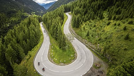 Serpentinenstraße, Alpenpass, Drohnenaufnahme, DJI-Drohne, Bergstraße, Luftaufnahme, Motorradtour, Kurvenfahrt, Motorradfahrer, Alpenlandschaft Die Luftaufnahme zeigt eine Serpentinen-Bergstraße in alpiner Landschaft, aufgenommen mit einer DJI-Drohne. Zwei Motorräder fahren durch eine enge Kehre, flankiert von dichten Nadelwäldern und steilen Hängen, im Hintergrund öffnet sich ein Tal. Die Perspektive aus der Höhe betont die geschwungene Straßenführung eines Alpenpasses und die Topografie.