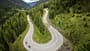 Die Luftaufnahme zeigt eine Serpentinen-Bergstraße in alpiner Landschaft, aufgenommen mit einer DJI-Drohne. Zwei Motorräder fahren durch eine enge Kehre, flankiert von dichten Nadelwäldern und steilen Hängen, im Hintergrund öffnet sich ein Tal. Die Perspektive aus der Höhe betont die geschwungene Straßenführung eines Alpenpasses und die Topografie.