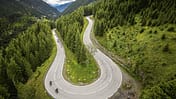Die Luftaufnahme zeigt eine Serpentinen-Bergstraße in alpiner Landschaft, aufgenommen mit einer DJI-Drohne. Zwei Motorräder fahren durch eine enge Kehre, flankiert von dichten Nadelwäldern und steilen Hängen, im Hintergrund öffnet sich ein Tal. Die Perspektive aus der Höhe betont die geschwungene Straßenführung eines Alpenpasses und die Topografie.