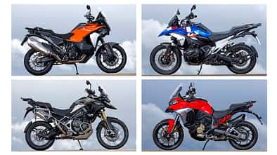 Vier Reiseenduro-Motorräder, BMW R 1300 GS Trophy, Ducati Multistrada V4 S, KTM 1390 Super Adventure S Evo und Triumph Tiger 1200 Rally Pro, fahren nebeneinander auf einem engen Weg. Die Straße führt von einem Aussichtspunkt neben einem Turm weg. Im Hintergrund ist eine weite Aussicht zu sehen. Alle vier Fahrer tragen Motorradbekleidung und Helme.