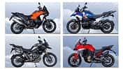 Vier Reiseenduro-Motorräder, BMW R 1300 GS Trophy, Ducati Multistrada V4 S, KTM 1390 Super Adventure S Evo und Triumph Tiger 1200 Rally Pro, fahren nebeneinander auf einem engen Weg. Die Straße führt von einem Aussichtspunkt neben einem Turm weg. Im Hintergrund ist eine weite Aussicht zu sehen. Alle vier Fahrer tragen Motorradbekleidung und Helme.