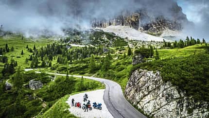 Motorrad, Alpen, Dolomiten, Bergstraße, Drohnenaufnahme, Motorradtour, Kurvenfahrt, Gebirge, Nebel, Landschaft Drohnenaufnahme einer kurvigen Bergstraße mit mehreren Reiseenduro-Motorrädern und Fahrern, umgeben von grüner Alpenlandschaft und Nebel, im Hintergrund ein steiler Felsabbruch.