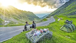 Das Foto zeigt eine Motorradreise in Rumänien mit Blick auf eine kurvige Bergpassstraße. Zwei Reisende in Motorradbekleidung machen am Straßenrand Rast; rechts im Bild stehen ein Zelt und eine beladene Reiseenduro, auf einem Fels liegen Proviant und Kochutensilien. Die Straße schlängelt sich durch eine grüne Hochgebirgslandschaft, während die tief stehende Sonne durch die Wolken bricht.