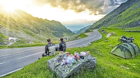 Das Foto zeigt eine Motorradreise in Rumänien mit Blick auf eine kurvige Bergpassstraße. Zwei Reisende in Motorradbekleidung machen am Straßenrand Rast; rechts im Bild stehen ein Zelt und eine beladene Reiseenduro, auf einem Fels liegen Proviant und Kochutensilien. Die Straße schlängelt sich durch eine grüne Hochgebirgslandschaft, während die tief stehende Sonne durch die Wolken bricht.