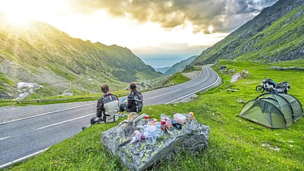 Motorradreise Rumänien, Gebirgsstraße Rumänien, Motorrad-Camping, Reiseenduro, Bergpass, Serpentinenstraße, Zelt, Berglandschaft, Sonnenuntergang, Motorrad-Reisefotografie Das Foto zeigt eine Motorradreise in Rumänien mit Blick auf eine kurvige Bergpassstraße. Zwei Reisende in Motorradbekleidung machen am Straßenrand Rast; rechts im Bild stehen ein Zelt und eine beladene Reiseenduro, auf einem Fels liegen Proviant und Kochutensilien. Die Straße schlängelt sich durch eine grüne Hochgebirgslandschaft, während die tief stehende Sonne durch die Wolken bricht.