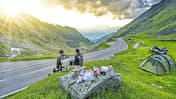 Das Foto zeigt eine Motorradreise in Rumänien mit Blick auf eine kurvige Bergpassstraße. Zwei Reisende in Motorradbekleidung machen am Straßenrand Rast; rechts im Bild stehen ein Zelt und eine beladene Reiseenduro, auf einem Fels liegen Proviant und Kochutensilien. Die Straße schlängelt sich durch eine grüne Hochgebirgslandschaft, während die tief stehende Sonne durch die Wolken bricht.