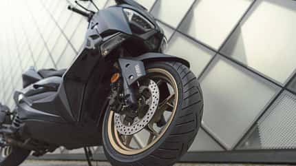 Continental Conti Scoot, Continental-Rollerreifen, Maxi-Scooter, Vorderreifen, Reifenprofil, Scheibenbremse, Leichtmetallfelge, Yamaha-Roller, Red Week, Detailaufnahme Das Foto zeigt den Rollerreifen Continental Conti Scoot an der Vorderachse eines Maxi-Scooters. In der Nahaufnahme sind das Reifenprofil, eine gelochte Scheibenbremse, der Bremssattel und eine goldfarbene Leichtmetallfelge zu erkennen; am Gabelschutz ist ein Yamaha‑Schriftzug sichtbar. Die Aufnahme steht im Zusammenhang mit der Continental‑Aktion Red Week (2026).