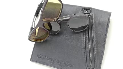 Motogoggles, Red Week, Motorradbrille, Motogoggles-Motorradbrille, Sonnenbrille, Motorrad-Zubehör, Brillenetui, Lederetui, Gelbfilter-Glas, Schutzbrille Die Aufnahme zeigt Motogoggles Motorradbrillen zusammen mit dem passenden Etui der Marke, dessen geprägter motogoggles‑Schriftzug sichtbar ist. Abgebildet sind zwei Brillen mit Kunststoffrahmen und getönten Gläsern, eine mit gelb getönten rechteckigen Scheiben und eine runde Sonnenbrille; das Etui besitzt einen seitlichen Reißverschluss. Studioaufnahme auf hellem Hintergrund.