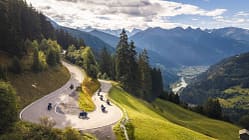 Das Luftbild zeigt eine Gruppe von Honda X‑Ride 2026 Motorrädern, die eine kurvige Serpentinenstraße in alpiner Berglandschaft befahren. Die erhöhte Perspektive betont die engen Kehren und den Verlauf der Passstraße, während im Hintergrund Tal, Flusslauf und Bergketten zu sehen sind. Die Aufnahme entstand per Drohne (DJI).