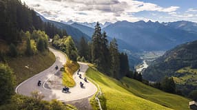 Das Luftbild zeigt eine Gruppe von Honda X‑Ride 2026 Motorrädern, die eine kurvige Serpentinenstraße in alpiner Berglandschaft befahren. Die erhöhte Perspektive betont die engen Kehren und den Verlauf der Passstraße, während im Hintergrund Tal, Flusslauf und Bergketten zu sehen sind. Die Aufnahme entstand per Drohne (DJI).