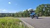 Naked-Bike, Straßenmotorrad, Kurvenfahrt, Schräglage, Landstraße, Motorradfahrer, Schutzkleidung, Helm, Sommerlandschaft, Straßenverkehr Fahrfoto eines Naked-Bikes auf der Landstraße, aufgenommen am 2026-07-21 laut Dateiname. Das blaue Straßenmotorrad wird in leichter Schräglage durch eine Rechtskurve gefahren; der Fahrer trägt Integralhelm, Handschuhe und Schutzkleidung. Im Hintergrund sind Bäume, Wiesen und Richtungstafeln unter klarem Sommerhimmel zu sehen.