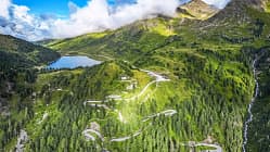 Die Drohnenaufnahme (DJI) zeigt eine alpine Berglandschaft mit einer serpentinenreichen Passstraße, dichten Nadelwäldern und einem hochgelegenen See. Rechts fällt ein Hang mit einem Wasserfall steil ab, während Wolken über den Gipfeln hängen und das weite Panorama teilweise beschatten. Der Straßenverlauf mit engen Kehren veranschaulicht die Topografie des Bergpasses.