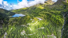 Die Drohnenaufnahme (DJI) zeigt eine alpine Berglandschaft mit einer serpentinenreichen Passstraße, dichten Nadelwäldern und einem hochgelegenen See. Rechts fällt ein Hang mit einem Wasserfall steil ab, während Wolken über den Gipfeln hängen und das weite Panorama teilweise beschatten. Der Straßenverlauf mit engen Kehren veranschaulicht die Topografie des Bergpasses.