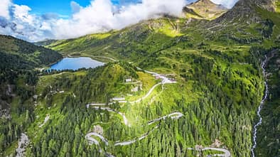 Die Drohnenaufnahme (DJI) zeigt eine alpine Berglandschaft mit einer serpentinenreichen Passstraße, dichten Nadelwäldern und einem hochgelegenen See. Rechts fällt ein Hang mit einem Wasserfall steil ab, während Wolken über den Gipfeln hängen und das weite Panorama teilweise beschatten. Der Straßenverlauf mit engen Kehren veranschaulicht die Topografie des Bergpasses.