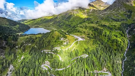 Luftaufnahme, Drohnenfoto, DJI-Drohne, Bergpass, Serpentinenstraße, Bergsee, Berglandschaft, Nadelwald, Wasserfall, Panorama Die Drohnenaufnahme (DJI) zeigt eine alpine Berglandschaft mit einer serpentinenreichen Passstraße, dichten Nadelwäldern und einem hochgelegenen See. Rechts fällt ein Hang mit einem Wasserfall steil ab, während Wolken über den Gipfeln hängen und das weite Panorama teilweise beschatten. Der Straßenverlauf mit engen Kehren veranschaulicht die Topografie des Bergpasses.