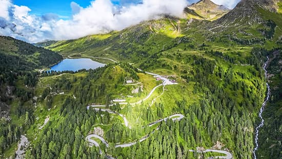 Die Drohnenaufnahme (DJI) zeigt eine alpine Berglandschaft mit einer serpentinenreichen Passstraße, dichten Nadelwäldern und einem hochgelegenen See. Rechts fällt ein Hang mit einem Wasserfall steil ab, während Wolken über den Gipfeln hängen und das weite Panorama teilweise beschatten. Der Straßenverlauf mit engen Kehren veranschaulicht die Topografie des Bergpasses.