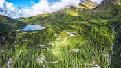 Die Drohnenaufnahme (DJI) zeigt eine alpine Berglandschaft mit einer serpentinenreichen Passstraße, dichten Nadelwäldern und einem hochgelegenen See. Rechts fällt ein Hang mit einem Wasserfall steil ab, während Wolken über den Gipfeln hängen und das weite Panorama teilweise beschatten. Der Straßenverlauf mit engen Kehren veranschaulicht die Topografie des Bergpasses.