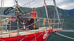 Ein Mann steht auf dem Deck eines roten Segelboots, das an einem Fjord liegt. Auf dem Vorschiff wird ein leichtes Motorrad (vermutlich ein Trial-Motorrad) mit einem Kran verladen oder gesichert. Im Hintergrund sind bewaldete Berghänge und ein Wasserfall zu sehen.