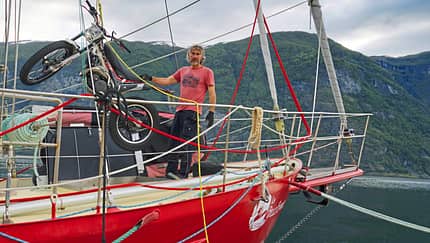 Ein Mann steht auf dem Deck eines roten Segelboots, das an einem Fjord liegt. Auf dem Vorschiff wird ein leichtes Motorrad (vermutlich ein Trial-Motorrad) mit einem Kran verladen oder gesichert. Im Hintergrund sind bewaldete Berghänge und ein Wasserfall zu sehen.
