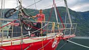 Ein Mann steht auf dem Deck eines roten Segelboots, das an einem Fjord liegt. Auf dem Vorschiff wird ein leichtes Motorrad (vermutlich ein Trial-Motorrad) mit einem Kran verladen oder gesichert. Im Hintergrund sind bewaldete Berghänge und ein Wasserfall zu sehen.