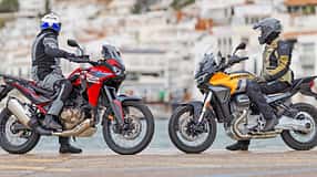 Das Foto zeigt eine Honda CRF1100L Africa Twin (links) und eine Moto Guzzi Stelvio (rechts) in Seitenansicht mit Fahrern in kompletter Schutzbekleidung. Beide Reiseenduros stehen am Ufer vor einer Küstenstadt; typische Adventure-Bike-Merkmale wie hoher Windschild, Speichenräder und aufrechte Sitzposition sind deutlich erkennbar.
