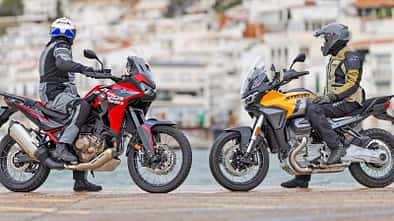 Das Foto zeigt eine Honda CRF1100L Africa Twin (links) und eine Moto Guzzi Stelvio (rechts) in Seitenansicht mit Fahrern in kompletter Schutzbekleidung. Beide Reiseenduros stehen am Ufer vor einer Küstenstadt; typische Adventure-Bike-Merkmale wie hoher Windschild, Speichenräder und aufrechte Sitzposition sind deutlich erkennbar.