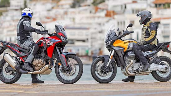 Das Foto zeigt eine Honda CRF1100L Africa Twin (links) und eine Moto Guzzi Stelvio (rechts) in Seitenansicht mit Fahrern in kompletter Schutzbekleidung. Beide Reiseenduros stehen am Ufer vor einer Küstenstadt; typische Adventure-Bike-Merkmale wie hoher Windschild, Speichenräder und aufrechte Sitzposition sind deutlich erkennbar.