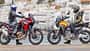Honda CRF1100L Africa Twin, Moto Guzzi Stelvio, Reiseenduro, Adventure-Bike, Motorradvergleich, Speichenrad, Windschild, Motorradtour, Küstenstadt, Motorradbekleidung Das Foto zeigt eine Honda CRF1100L Africa Twin (links) und eine Moto Guzzi Stelvio (rechts) in Seitenansicht mit Fahrern in kompletter Schutzbekleidung. Beide Reiseenduros stehen am Ufer vor einer Küstenstadt; typische Adventure-Bike-Merkmale wie hoher Windschild, Speichenräder und aufrechte Sitzposition sind deutlich erkennbar.