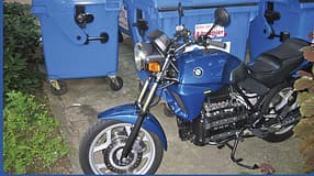 Blaues BMW-Motorrad vor blauen Müllcontainern im Freien, teilweise von Pflanzen umgeben.