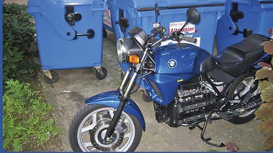 Blaues BMW-Motorrad vor blauen Müllcontainern im Freien, teilweise von Pflanzen umgeben.