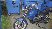 BMW-Motorrad, Straßenmotorrad, Motorrad, Papiercontainer, Stadtumgebung, Altpapier, Zweirad, Parken, BMW Blaues BMW-Motorrad vor blauen Müllcontainern im Freien, teilweise von Pflanzen umgeben.