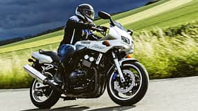 Yamaha Fazer 600 fährt in Kurvenfahrt auf Landstraße. Hintergrund eine ländliche Umgebung mit grünen Wiesen und ein dunkel bewölkter Himmel.