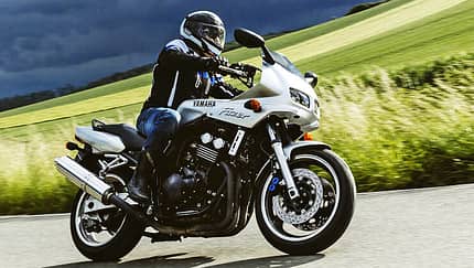 Yamaha Fazer 600, Landstraße, Kurvenfahrt, Straßenmotorrad, Fahrer, Sommerlandschaft Yamaha Fazer 600 fährt in Kurvenfahrt auf Landstraße. Hintergrund eine ländliche Umgebung mit grünen Wiesen und ein dunkel bewölkter Himmel.