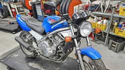 Honda CB-1 in blauer Lackierung auf einer Werkstattbühne, umgeben von Werkstattregalen und Werkzeugen.