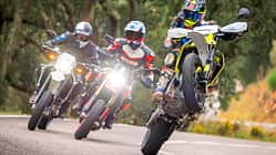 Drei Supermoto-Motorräder, Ducati Hypermotard 698 Mono RVE, Husqvarna 701 Supermoto und Suzuki DR-Z4SM, fahren auf einer Landstraße. Der Fahrer auf der Husqvarna 701 Supermoto macht einen Wheelie, die anderen beiden fahren nebeneinander. Alle Fahrer tragen Motorradschutzkleidung und Helme. Die Szene spielt sich auf einer kurvigen Straße im Grünen ab.