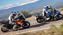 Zwei Reiseenduro-Motorräder, BMW R 1300 GS Trophy und KTM 1390 Super Adventure S Evo, fahren nebeneinander in Kurvenfahrt auf einem Schotterweg. Beide Fahrer tragen Motorradbekleidung und Helme. Im Hintergrund sind Bäume, Steine und Berge zu sehen.
