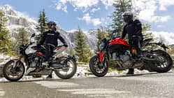 Ducati Monster+, Triumph Trident 800, Alpen Masters 2026, Mid-Naked Bikes, Alpen, Motorradtest, Naked Bike Zwei Mid-Naked-Motorräder mit Fahrern auf Alpenstraße, Schneereste am Straßenrand, Gebirgslandschaft im Hintergrund.
