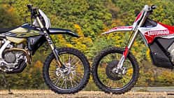 Enduro, Motorrad, Geländemotorrad, Offroad, Motorradtechnik, Windenergie, Wald, Motorsport, Zweirad, Natur, Triumph TF 450-E, Stark Varg EX Zwei Enduro-Motorräder, die Stark Varg EX und die Triumph TF 450-E parken frontal gegenüber im Gelände vor bewaldetem Hintergrund und Windrädern, sodass sich die Vorderreifen berühren. Aufnahme aus der Seitenansicht.