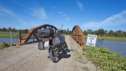 Reiseenduro, Adventure-Bike, Motorrad, Seitenkoffer, Topcase, Stollenreifen, Schotterstraße, Stahlbrücke, Gewichtsbeschränkung, Flusslandschaft Das Foto zeigt ein Reiseenduro‑Motorrad mit montierten Seitenkoffern und Topcase auf einer Schotterpiste vor einer kleinen stählernen Brücke über ein Gewässer. Stollenreifen, hohe Bodenfreiheit und Gepäcksystem betonen den Offroad‑Einsatz. Am Brückeneingang steht ein Schild mit der Aufschrift „WEIGHT LIMIT 5 TONS“ in ländlicher Umgebung unter blauem Himmel.