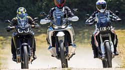 Die Aufnahme zeigt drei Reiseenduros in der Frontansicht: CFMOTO 1000MT-X, Ducati DesertX und Honda CRF1100L Africa Twin. Die Adventure-Bikes fahren nebeneinander auf einer Schotterstraße, mit hohen Windschilden und Handprotektoren gut erkennbar. Im leichten Offroad-Einsatz wirbeln sie Staub auf, die Szene wirkt wie ein Vergleichslauf im Gelände.