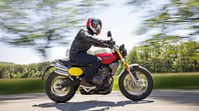 Fantic Caballero 700 Motorrad in Fahrt auf Landstraße, seitliche Ansicht, Fahrer mit Helm und Lederjacke, unscharfer Hintergrund durch Mitzieheffekt.