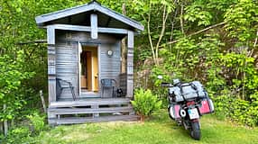 Reiseenduro Moto Guzzi V85 TT mit Gepäck vor einer kleinen Holzhütte im Grünen. Die Szene zeigt eine typische Motorradtour-Unterkunft.