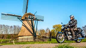 Motorradfahrer auf Landstraße vor Windmühle und Windkraftanlage, ländliche Umgebung, blauer Himmel.