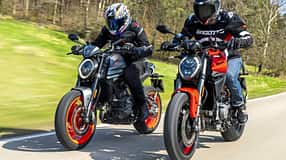 Zwei Ducati Monster Motorräder, neu und alt, fahren nebeneinander auf einer Landstraße. Beide Fahrer tragen Motorradbekleidung und Helme. Die Aufnahme zeigt einen direkten Vergleich der beiden Modellgenerationen in Bewegung.