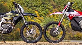 Zwei Enduro-Motorräder, die Stark Varg EX und die Triumph TF 450-E parken frontal gegenüber im Gelände vor bewaldetem Hintergrund und Windrädern, sodass sich die Vorderreifen berühren. Aufnahme aus der Seitenansicht.