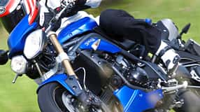 Eine Triumph Speed Triple in blauer Lackierung wird in Schräglage auf einer Landstraße gefahren. Der Fahrer trägt schwarze Motorradbekleidung und einen rot-weißen Helm.